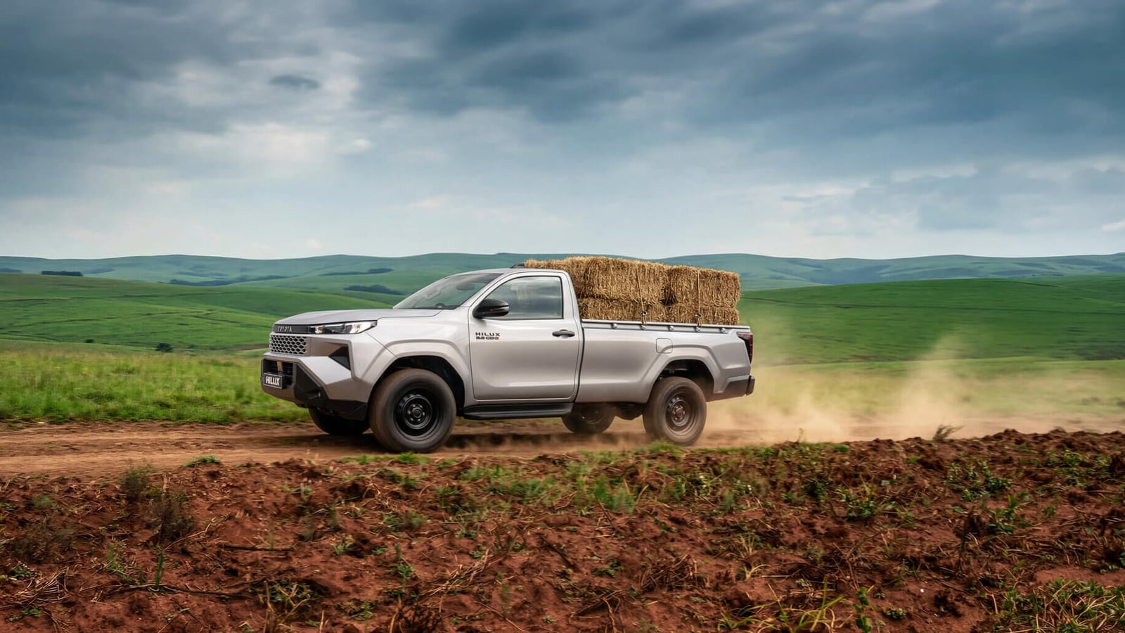 A Toyota Hilux single cab carrying hay bales along a dirt road through green farmland