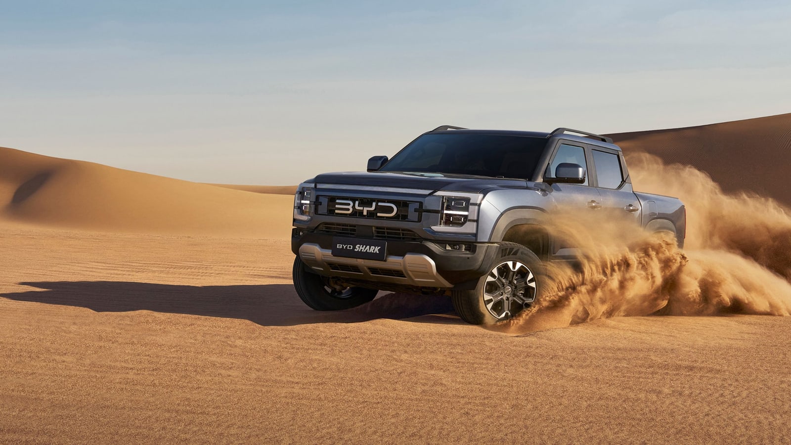 A silver BYD Shark 6 double cab throwing a plume of sand as it powers across desert dunes at low sun