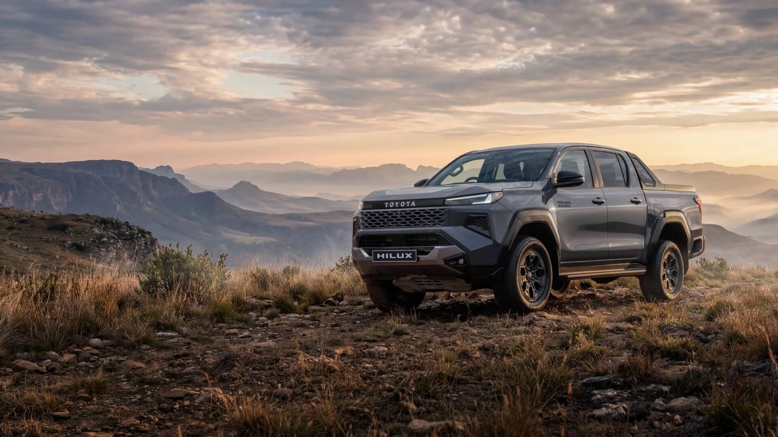 A grey new-generation Toyota Hilux double cab parked on a rocky ridge with a misty escarpment behind it at sunrise
