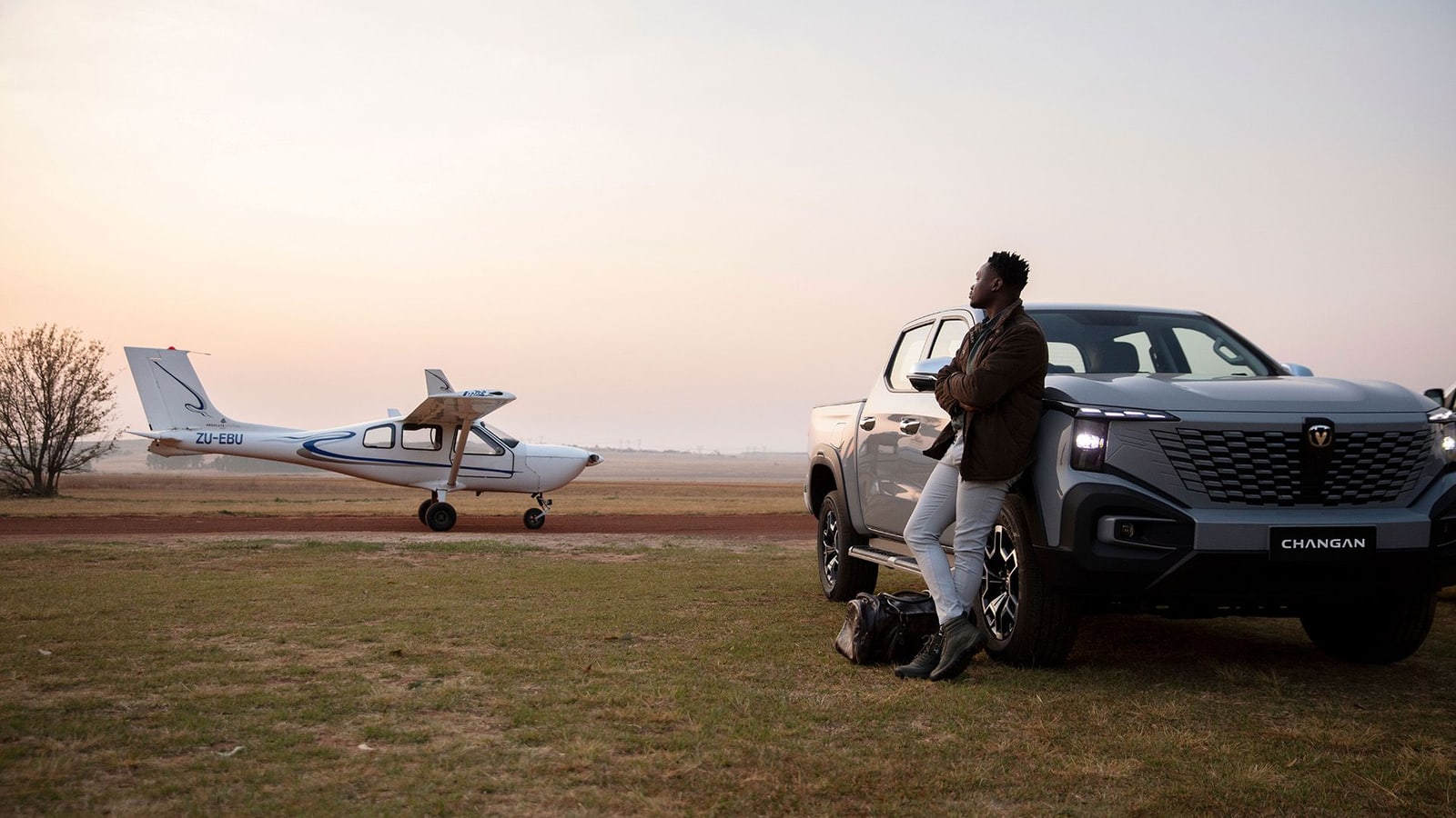 A grey Changan Hunter Reev double cab on a grass airstrip at dusk with a light aircraft taxiing behind it