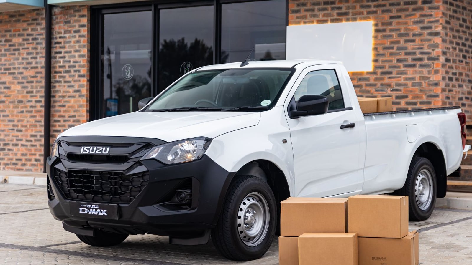 A white Isuzu D-Max single cab on steel wheels parked at an office block with cardboard boxes stacked beside the load bin