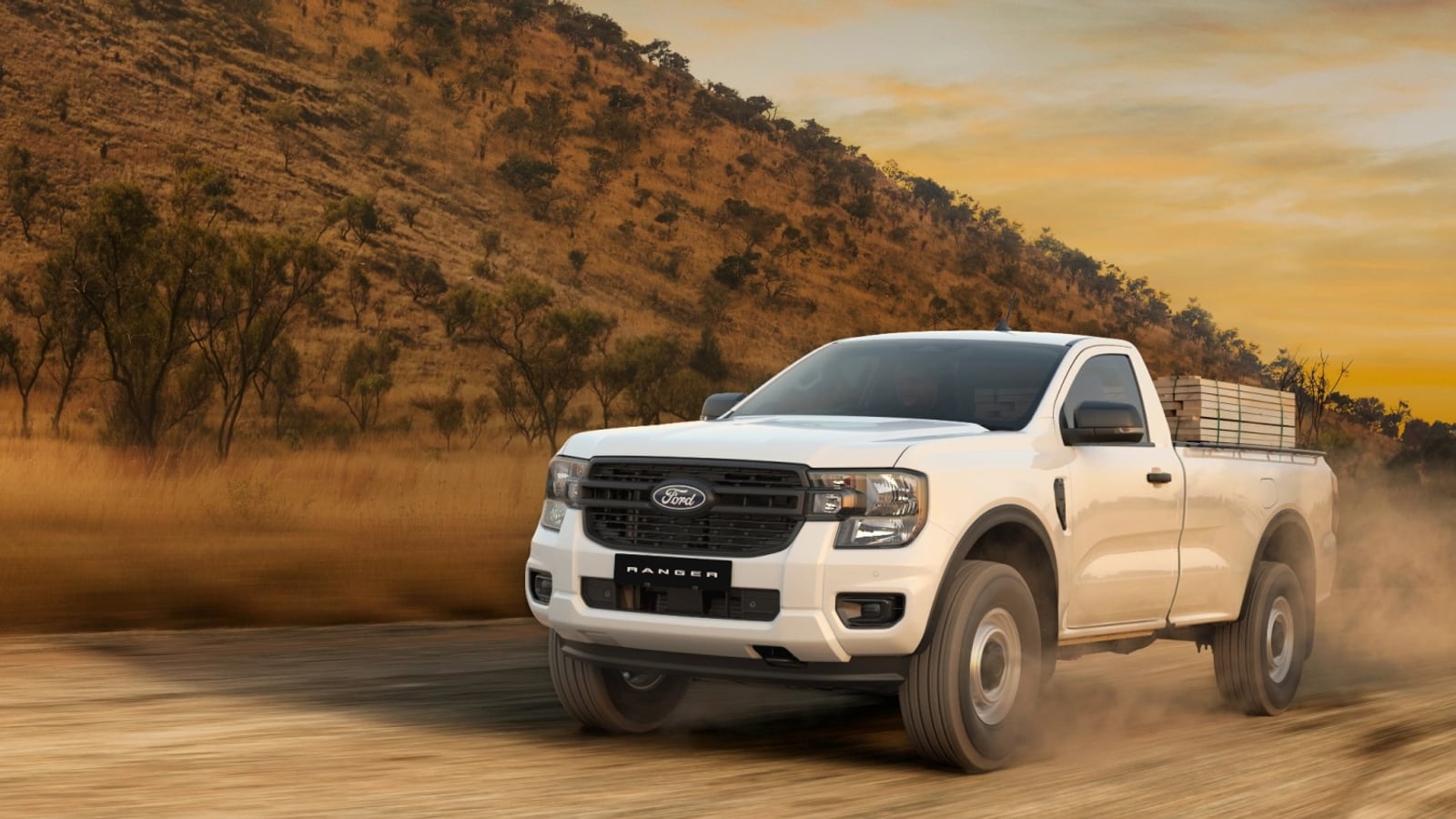 A white Ford Ranger XL single cab on steel wheels with a load of timber, on a dust road at sunset