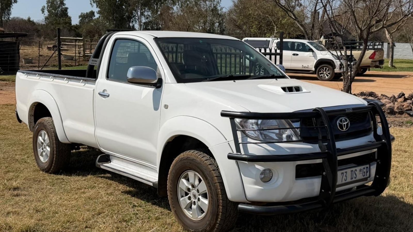 A white 2011 Toyota Hilux 3.0 D-4D Raider single cab with a bullbar and the bonnet scoop, parked on a plot with a farm gate and bluegum trees behind it
