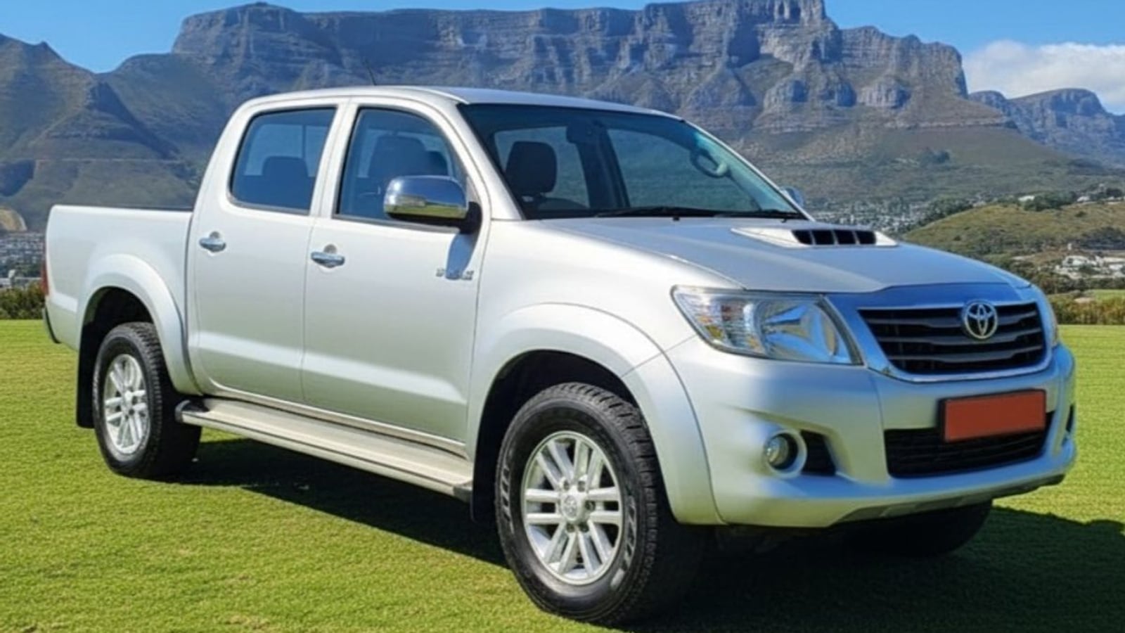 A silver 2014 Toyota Hilux 3.0 D-4D Raider double cab parked on grass with Table Mountain behind it