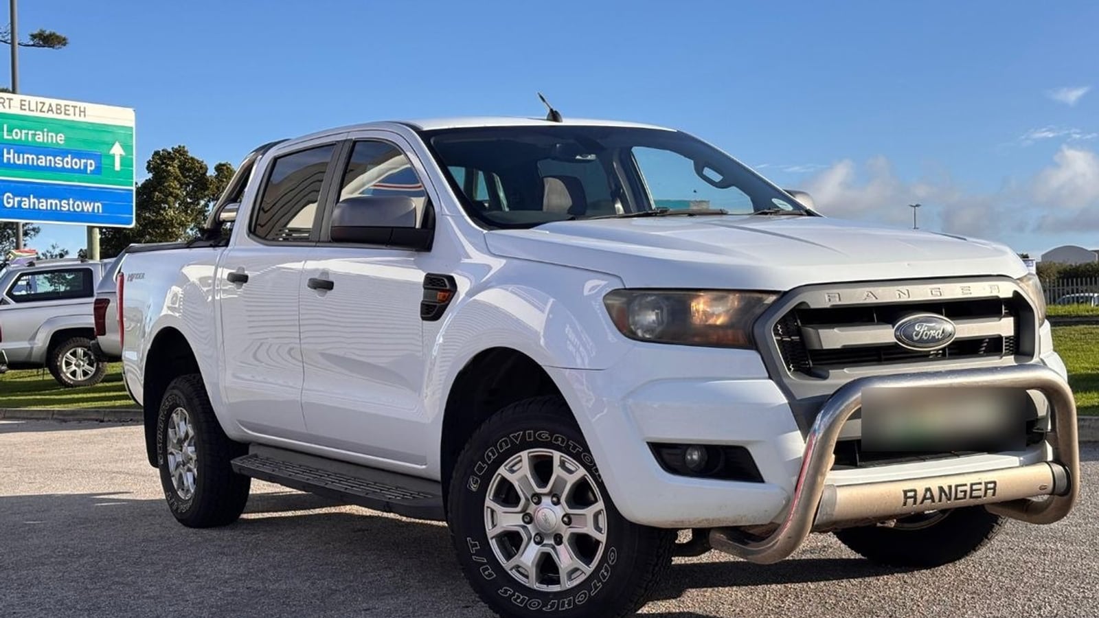 A white 2016 Ford Ranger 2.2 TDCi XLS double cab Hi-Rider with a nudge bar and all-terrain tyres, parked in Gqeberha with an N2 road sign for Humansdorp and Grahamstown behind it