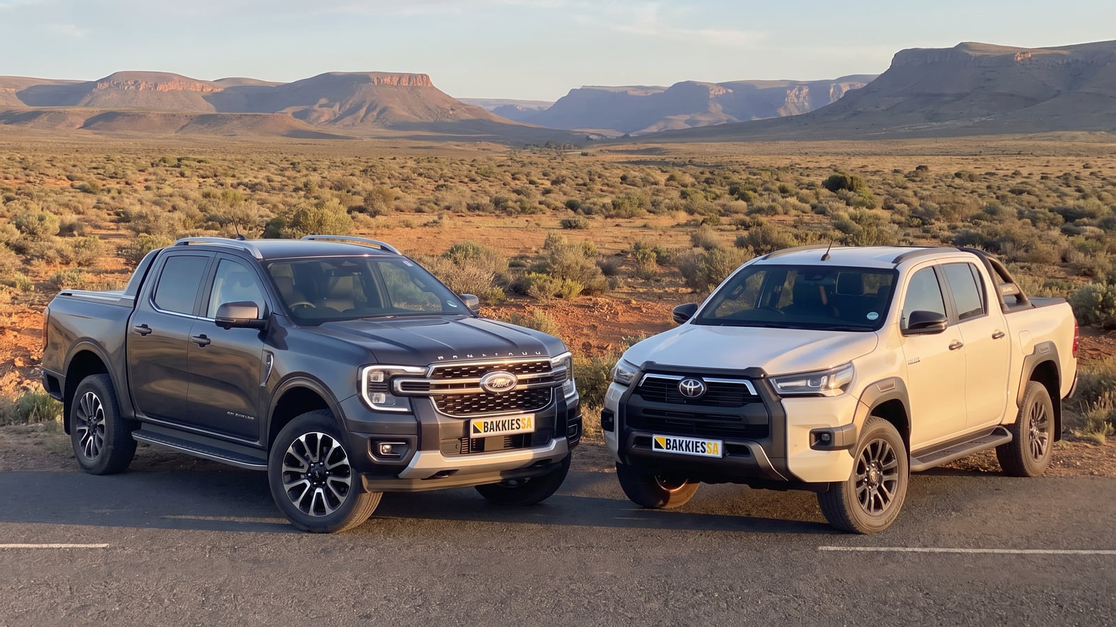 A Ford Ranger and a Toyota Hilux double cab parked side by side on a Karoo road at sunset