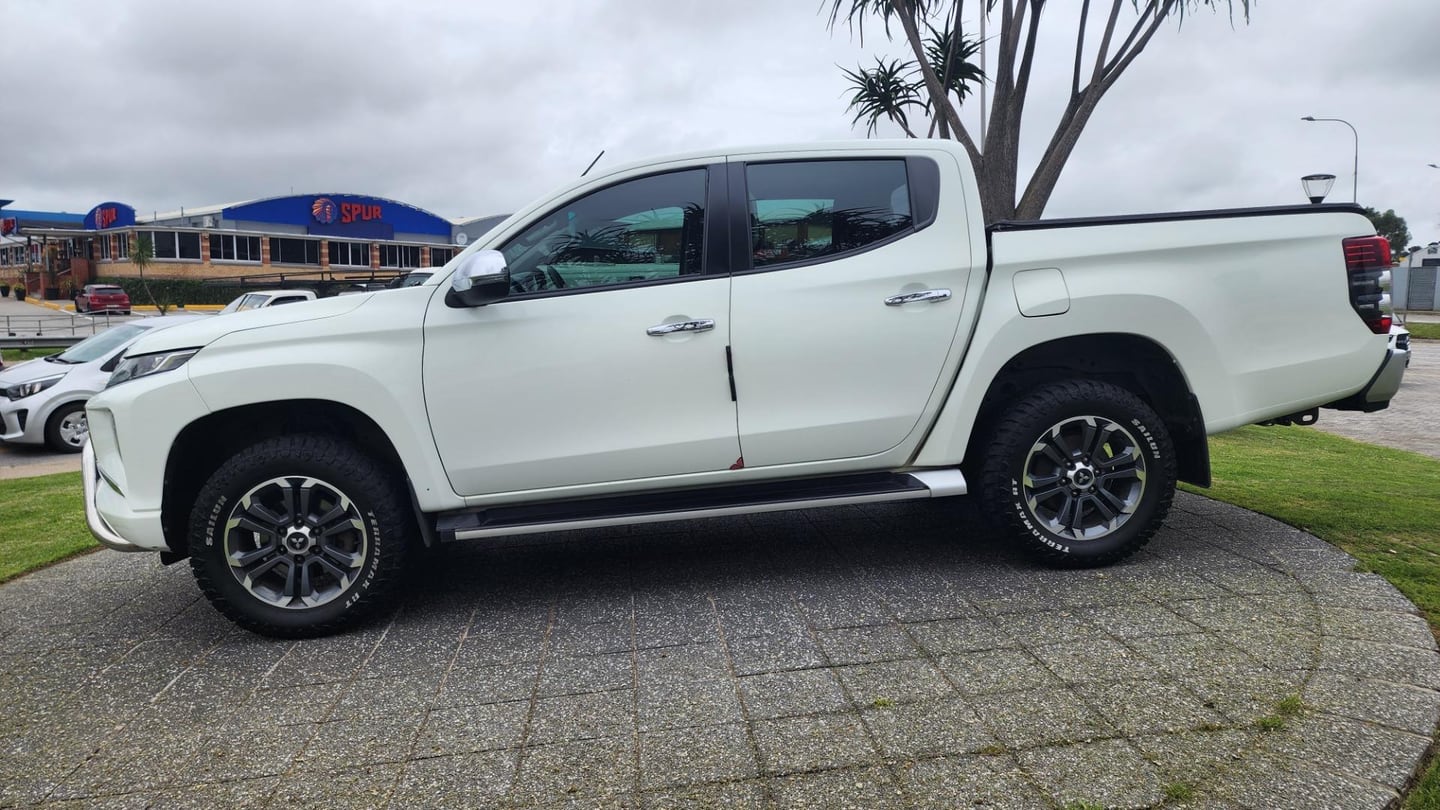 A white 2020 Mitsubishi Triton 2.4 DI-D automatic double cab with black alloys and a roll bar, side on, on paving beside a lawn in Port Elizabeth