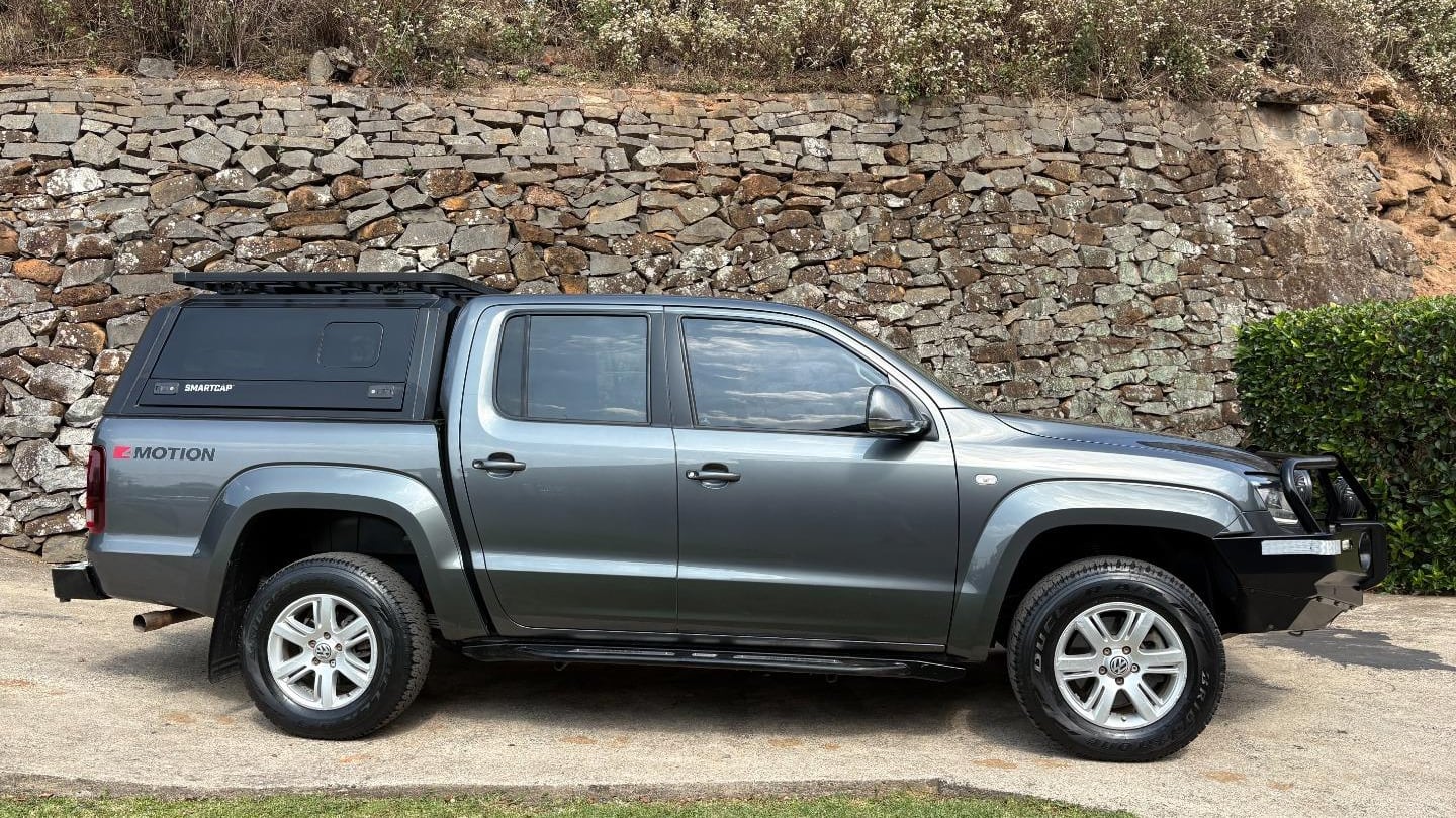 A grey 2021 Volkswagen Amarok 3.0 V6 Highline 4Motion double cab with a black canopy, roof rack and bull bar, side on, parked on a lawn below a dry-stone wall