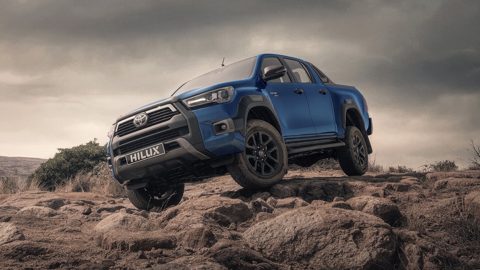 A blue Toyota Hilux 2.8 GD-6 Legend double cab climbing a rocky slope under a storm sky