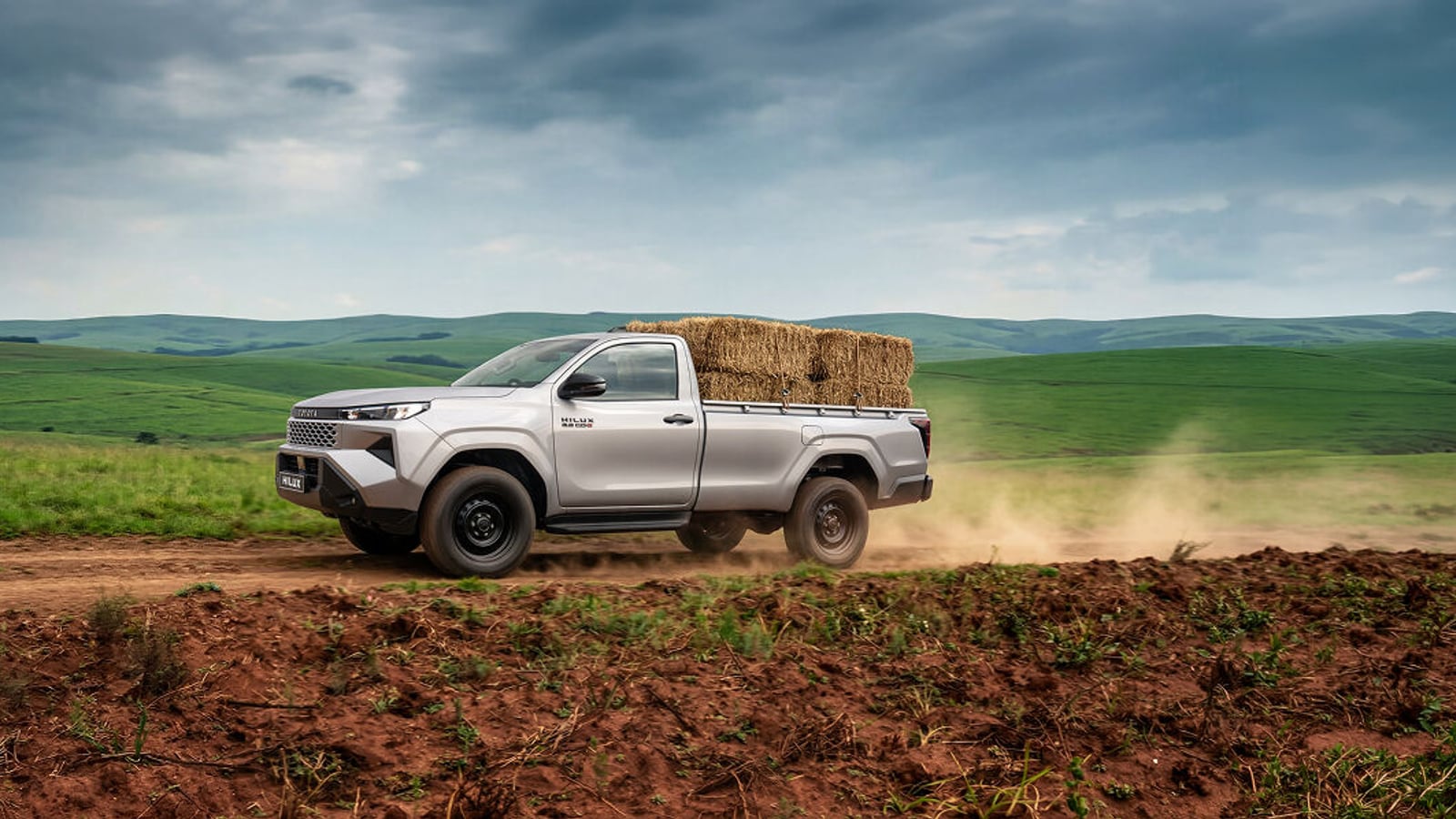 A Toyota Hilux single cab carrying hay bales along a dirt road through green farmland