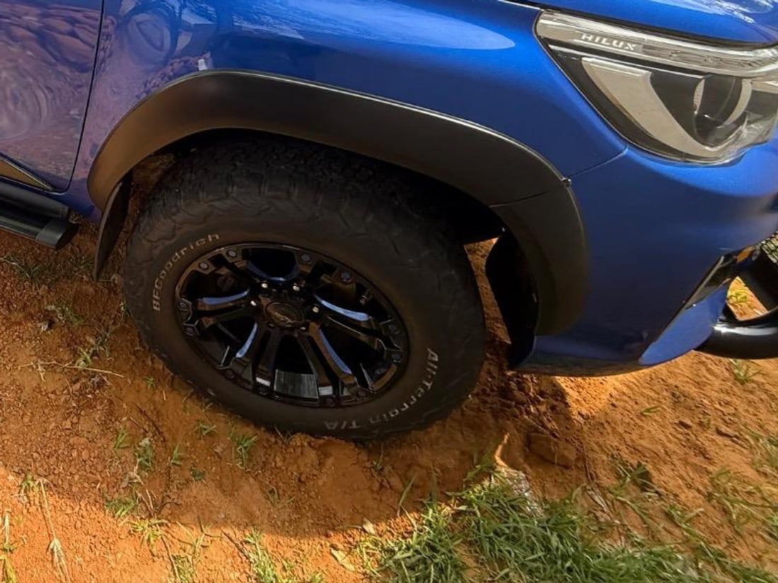 A close-up of a BF Goodrich All-Terrain tyre on a black alloy wheel, sitting in loose red soil, with the brand name readable on the sidewall