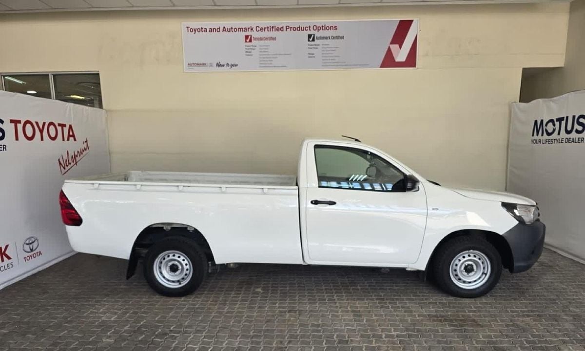 A white 2021 Toyota Hilux 2.0 VVTi single cab seen from the side, on silver steel wheels with tall narrow tyres, parked on brick paving at a dealer in Nelspruit