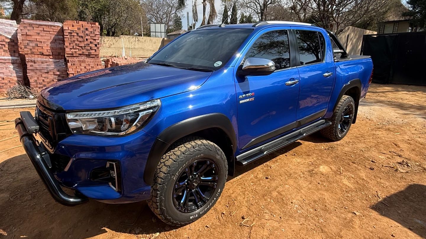 A blue 2021 Toyota Hilux Legend double cab with a small suspension lift and black wheels with all-terrain tyres, parked on red soil beside a half-built brick building in Gauteng