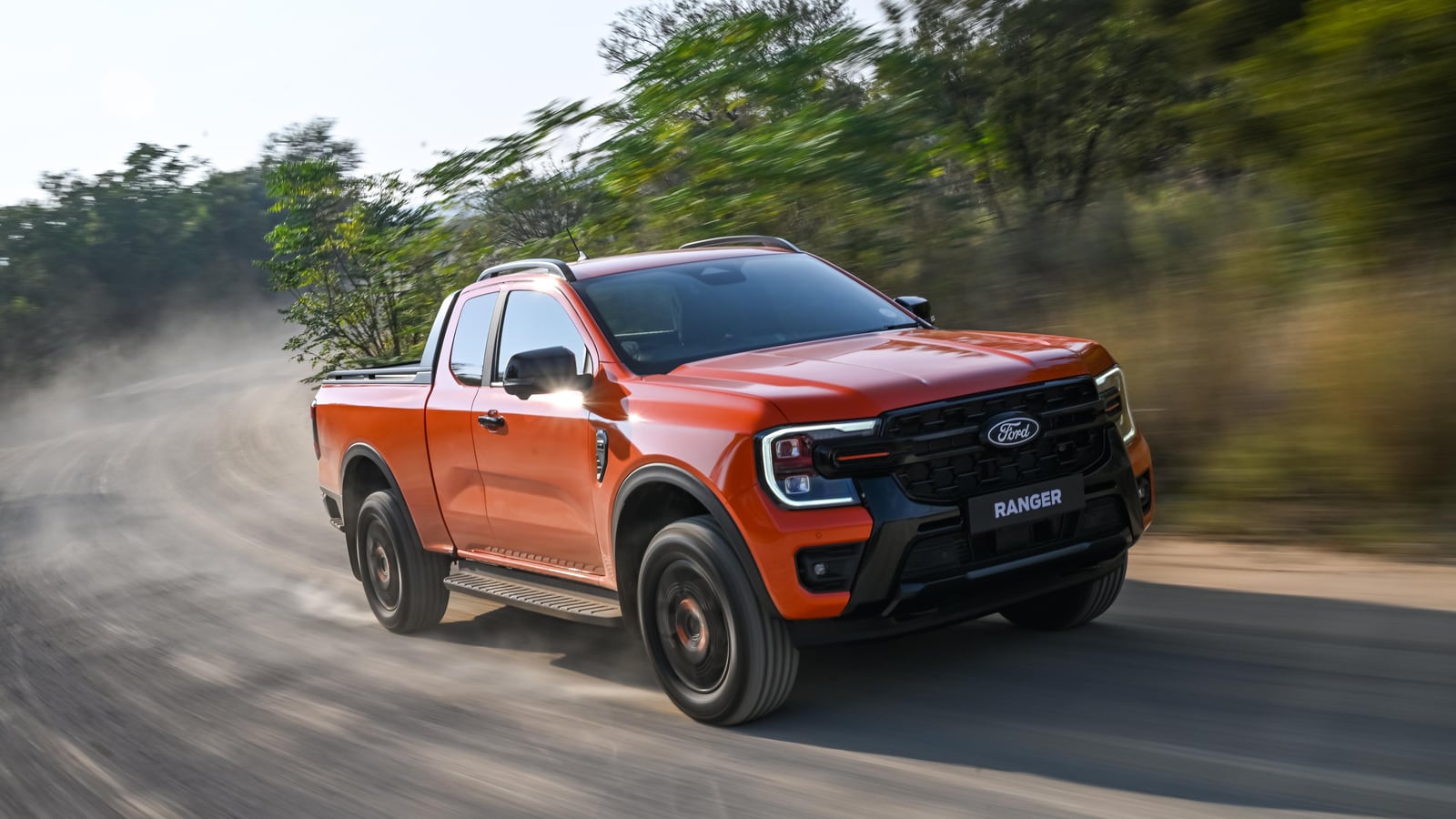 An orange Ford Ranger Wildtrak driving fast along a dirt road in South Africa with a thick plume of dust behind it and bush on either side